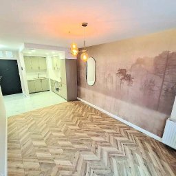 APARTMENTS REAL ESTATE SPÓŁKA Z OGRANICZONĄ ODPOWIEDZIALNOŚCIĄ - Wykończone mieszkanie z parkietem ułożonym w jodełkę, fototapetą z motywem leśnym i nowoczesnym oświetleniem nad stołem. Widoczny fragment kuchni z jasnymi szafkami.