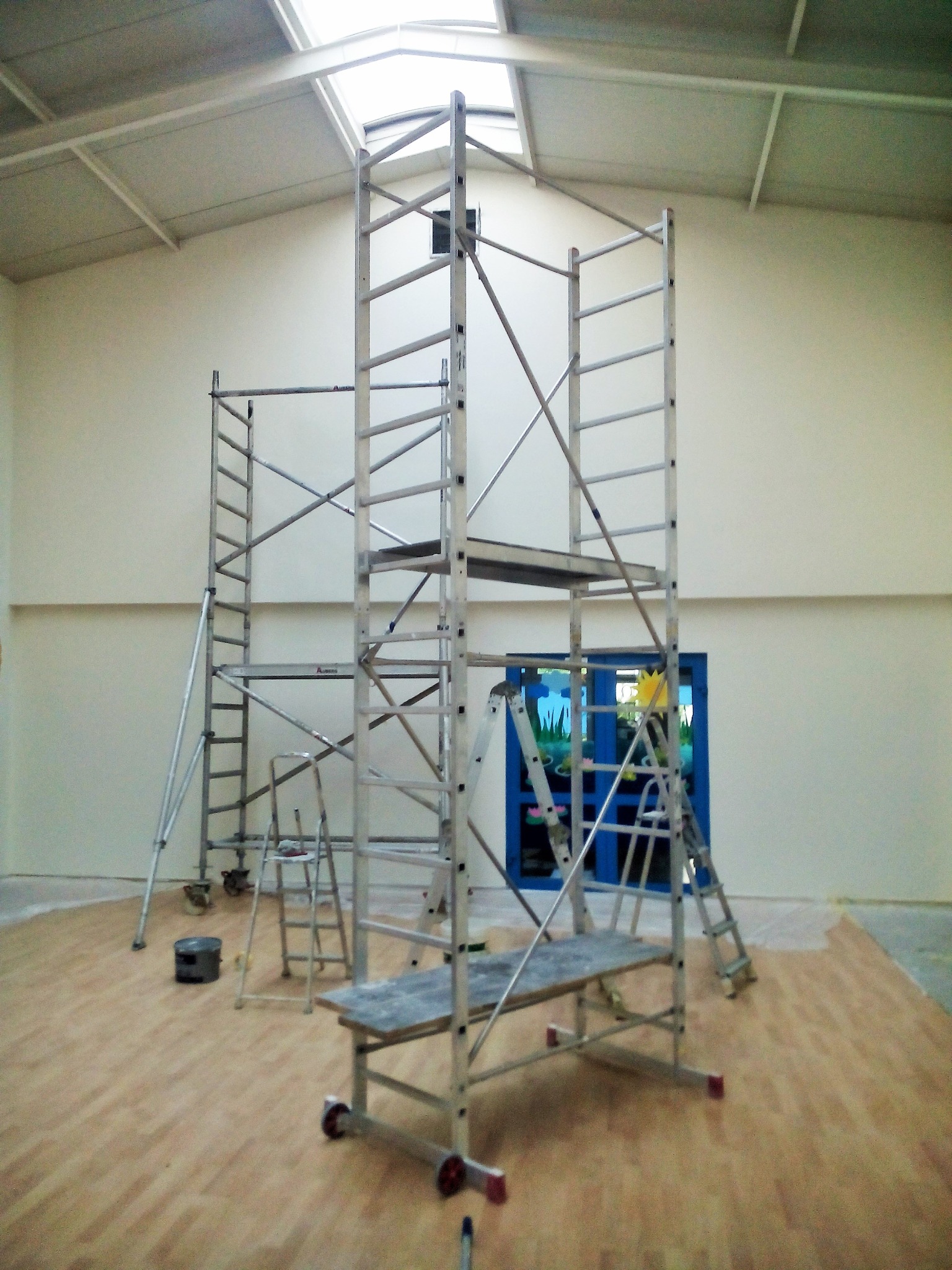 Wnętrze hali z drewnopodobną podłogą i białymi ścianami, na których rozstawione są aluminiowe rusztowania, a w tle widoczne niebieskie drzwi z dziecięcymi malunkami.