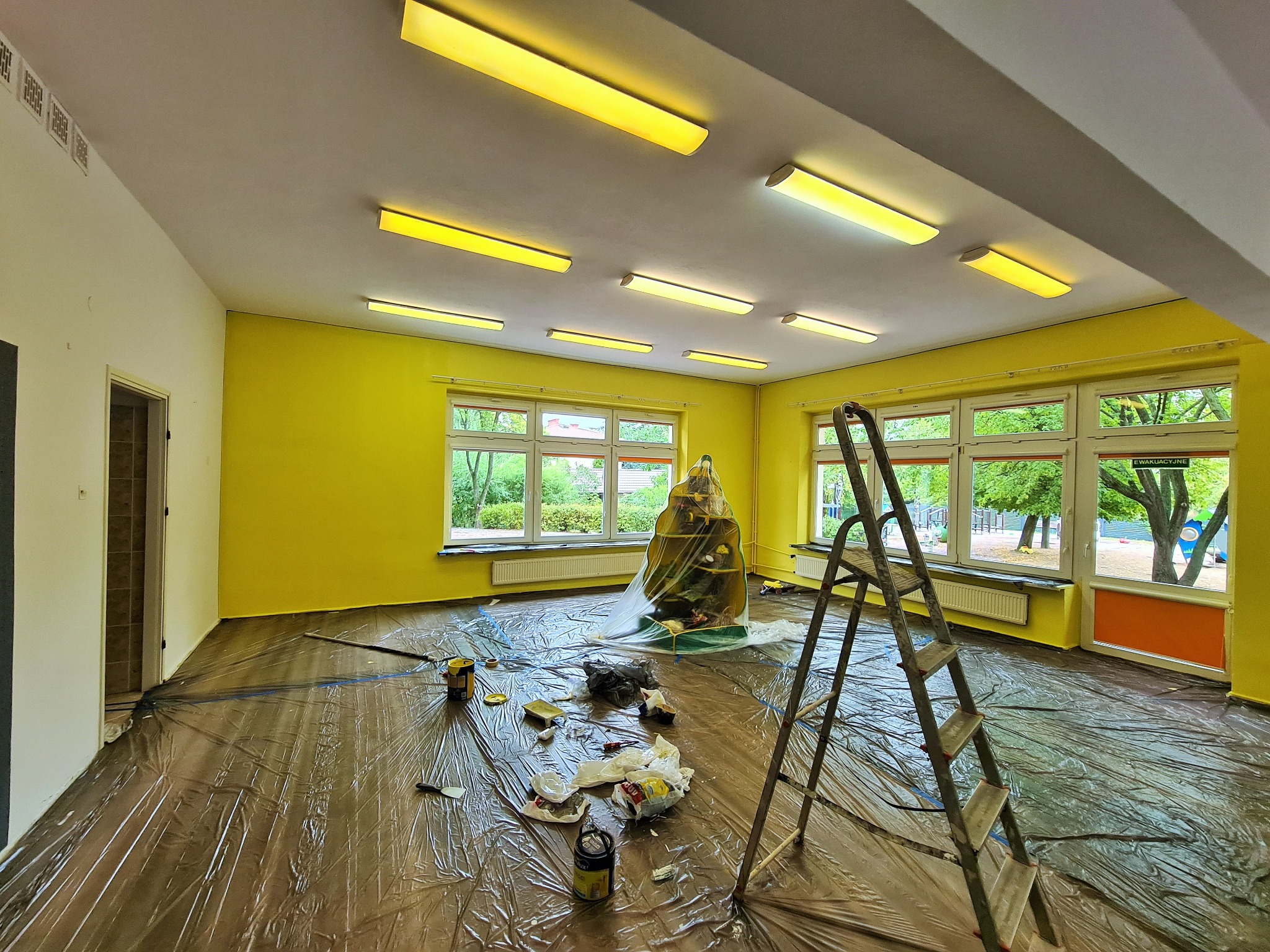 Sala przedszkolna w trakcie malowania na żółto, podłoga zabezpieczona folią, widoczna drabina i puszki z farbą.