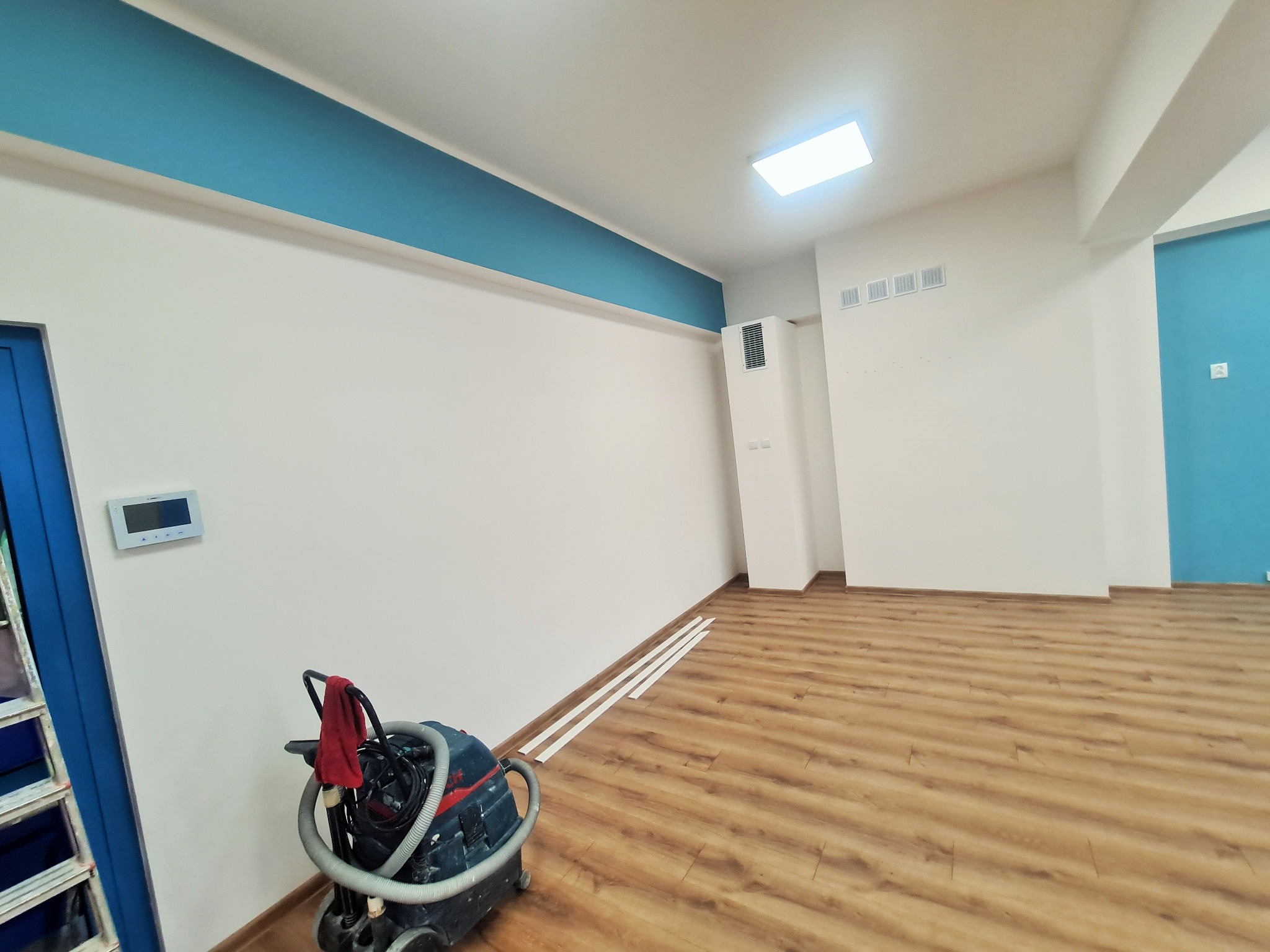 Wnętrze biura po malowaniu ścian i sufitów, widoczny odkurzacz przemysłowy, listwy przypodłogowe i świeżo pomalowane ściany w kolorze białym i turkusowym.