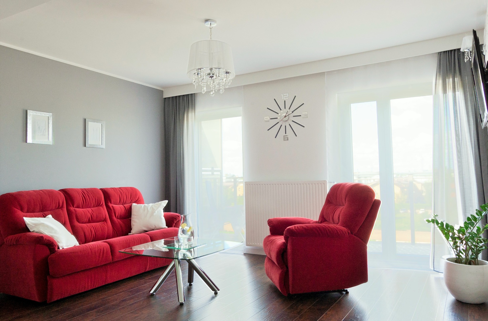 Jasne wnętrze salonu z ciemną drewnianą podłogą, czerwonym welurowym kompletem wypoczynkowym, szarą ścianą z ramkami i dużym oknem z firanami.