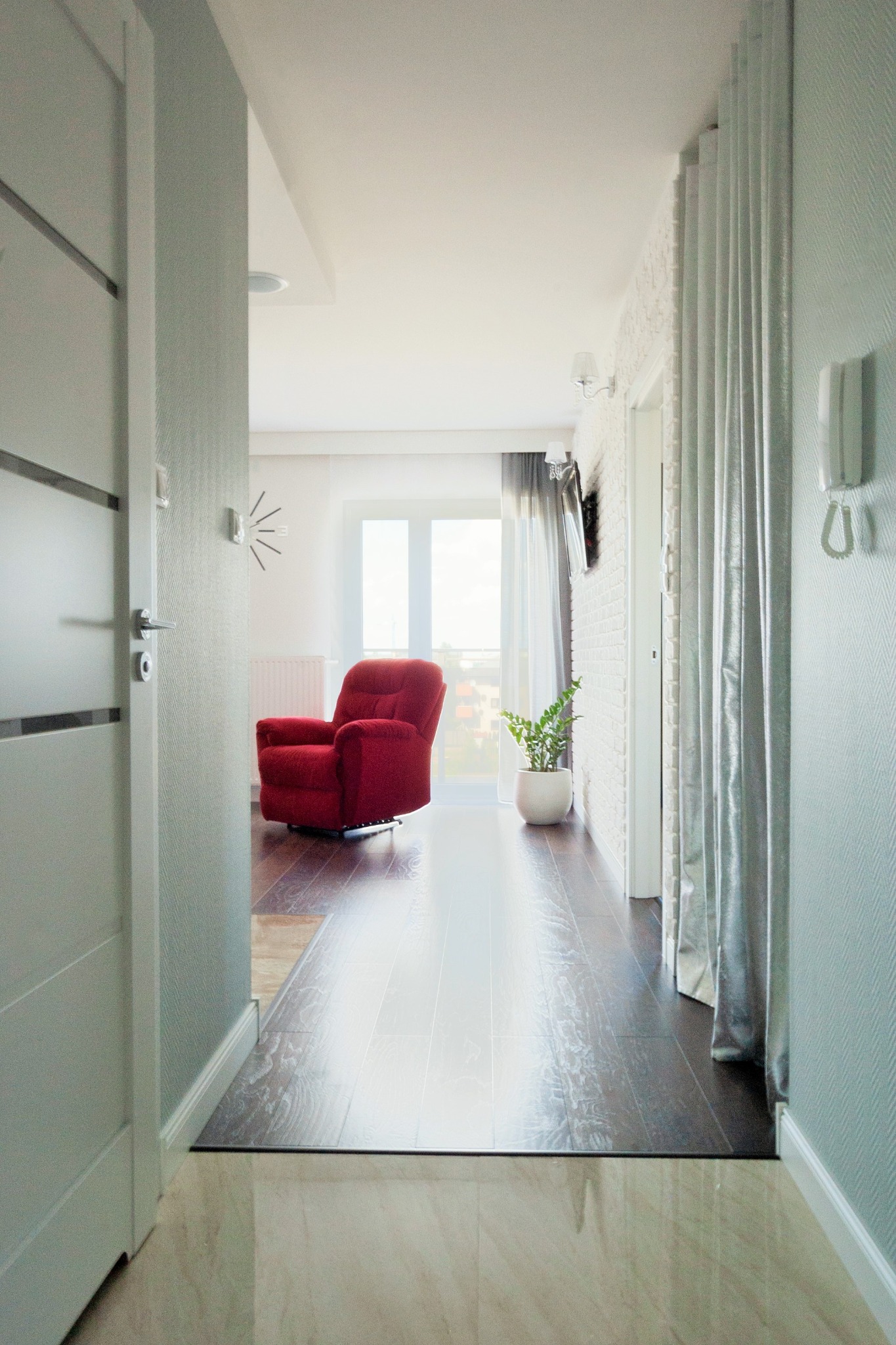 Widok z korytarza na salon: czerwony fotel relaksacyjny na ciemnej podłodze z paneli, białe ściany z cegły, roślina doniczkowa, okno z widokiem na miasto, jasne zasłony i szara ściana z tapetą...