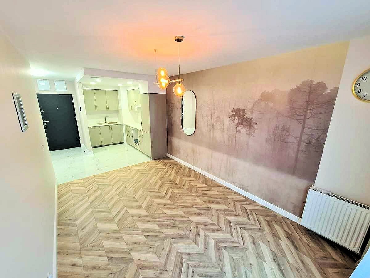 Wykończone mieszkanie z parkietem ułożonym w jodełkę, fototapetą z motywem leśnym i nowoczesnym oświetleniem nad stołem. Widoczny fragment kuchni z jasnymi szafkami.