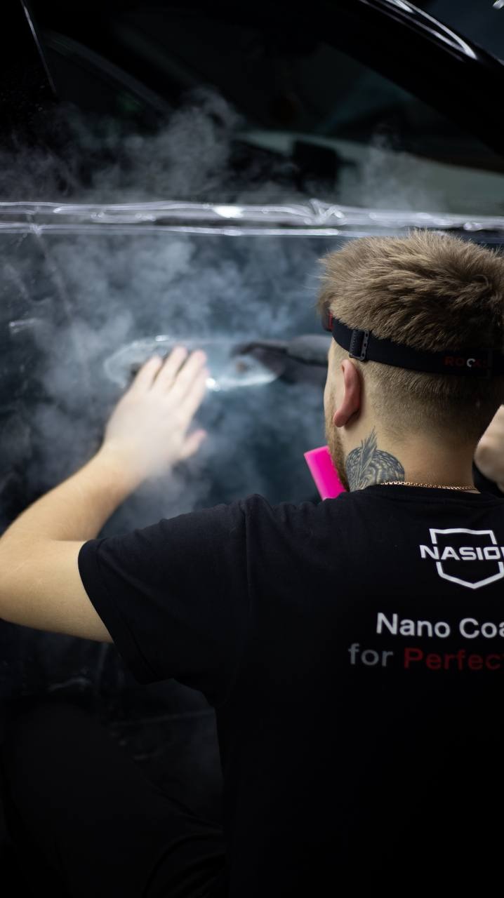 Pracownik w czarnej koszulce z logo firmy aplikuje folię ochronną na szybę samochodu, widoczna para wodna.