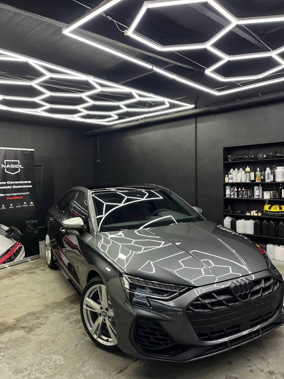 Szary samochód Audi w studiu detailingowym z heksagonalnym oświetleniem sufitowym, widoczny plakat reklamowy Nasioł i półki z kosmetykami samochodowymi.