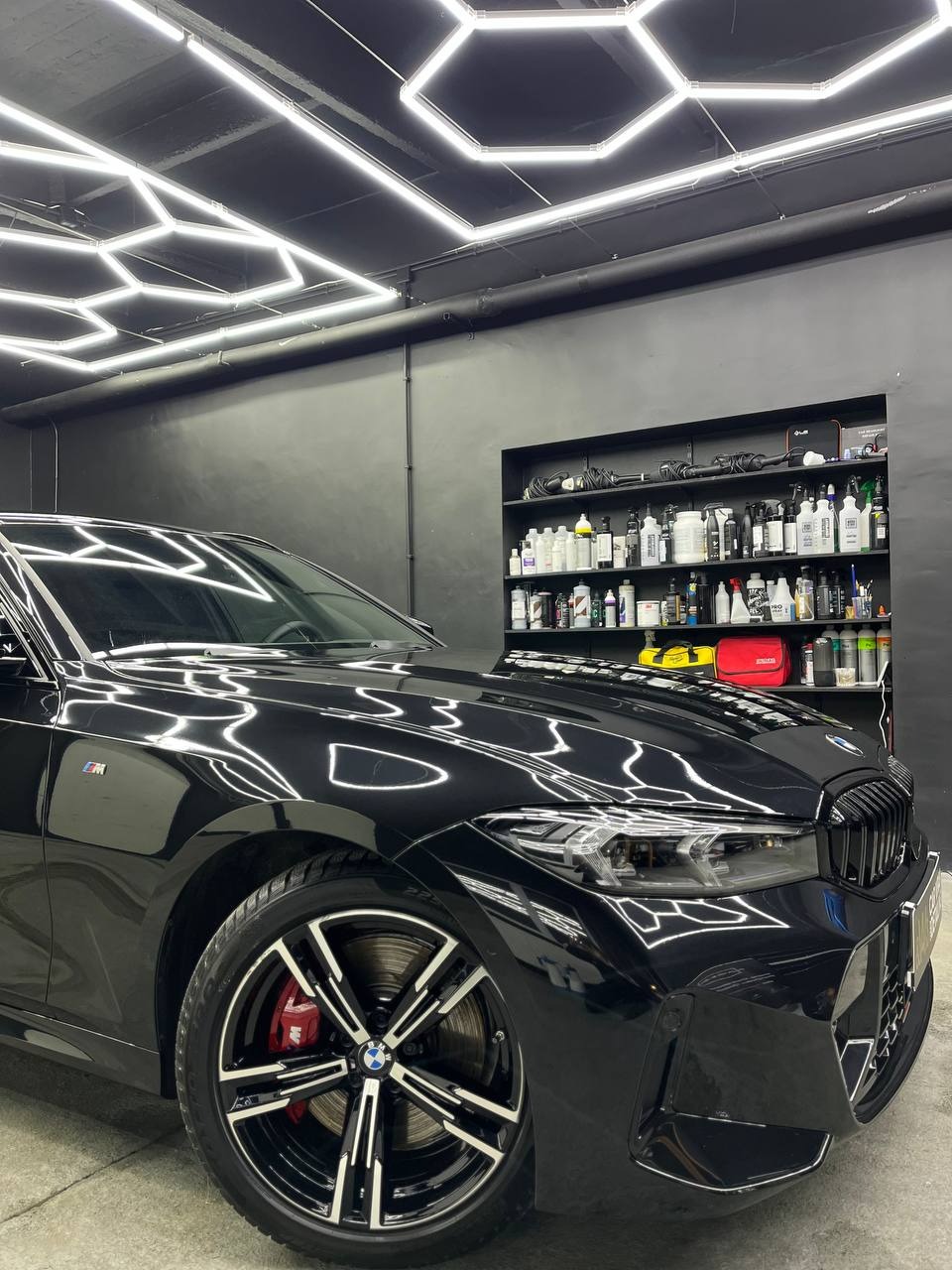 Błyszczący czarny samochód BMW po detailingu, widoczne odbicia światła LED na karoserii, czerwone zaciski hamulcowe, tło stanowi ściana z półkami pełnymi preparatów do pielęgnacji aut.