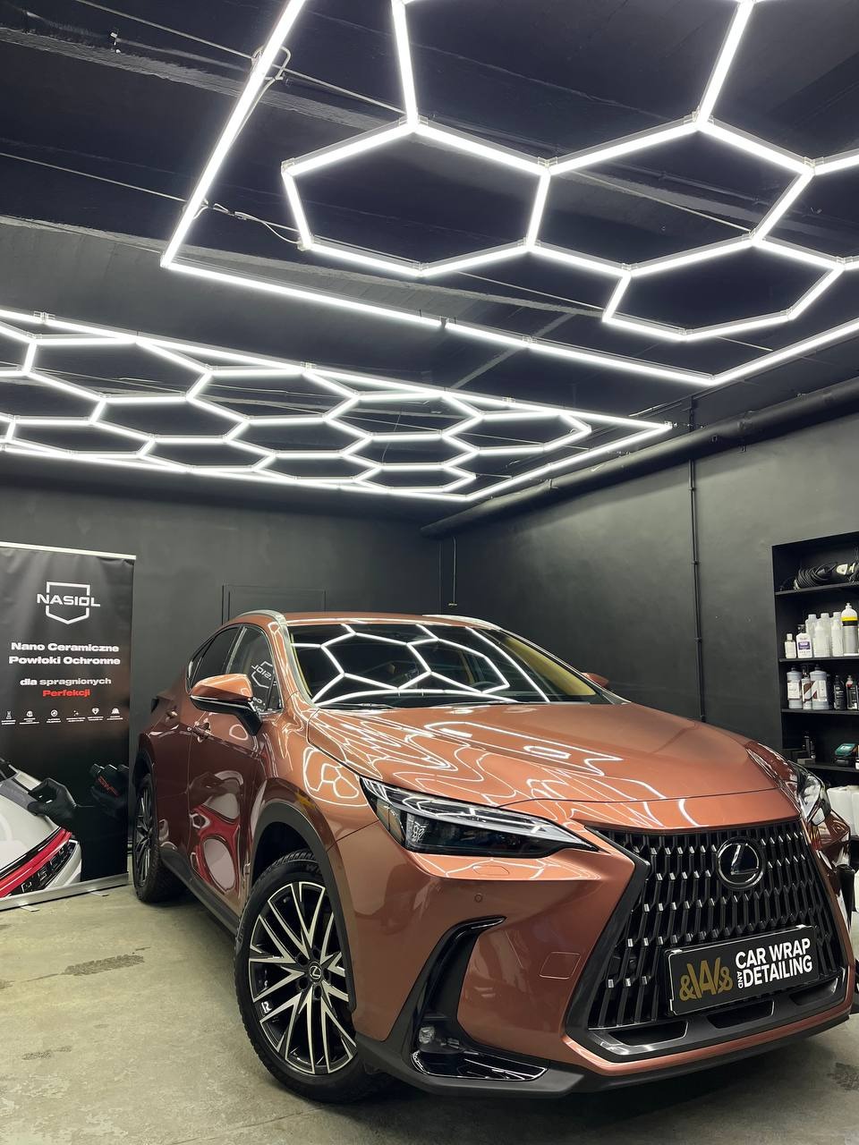 Brązowy Lexus NX po aplikacji folii ochronnej, widoczne logo 'A&A Car Wrap Detailing' na atrapie chłodnicy, oświetlenie heksagonalne na suficie.
