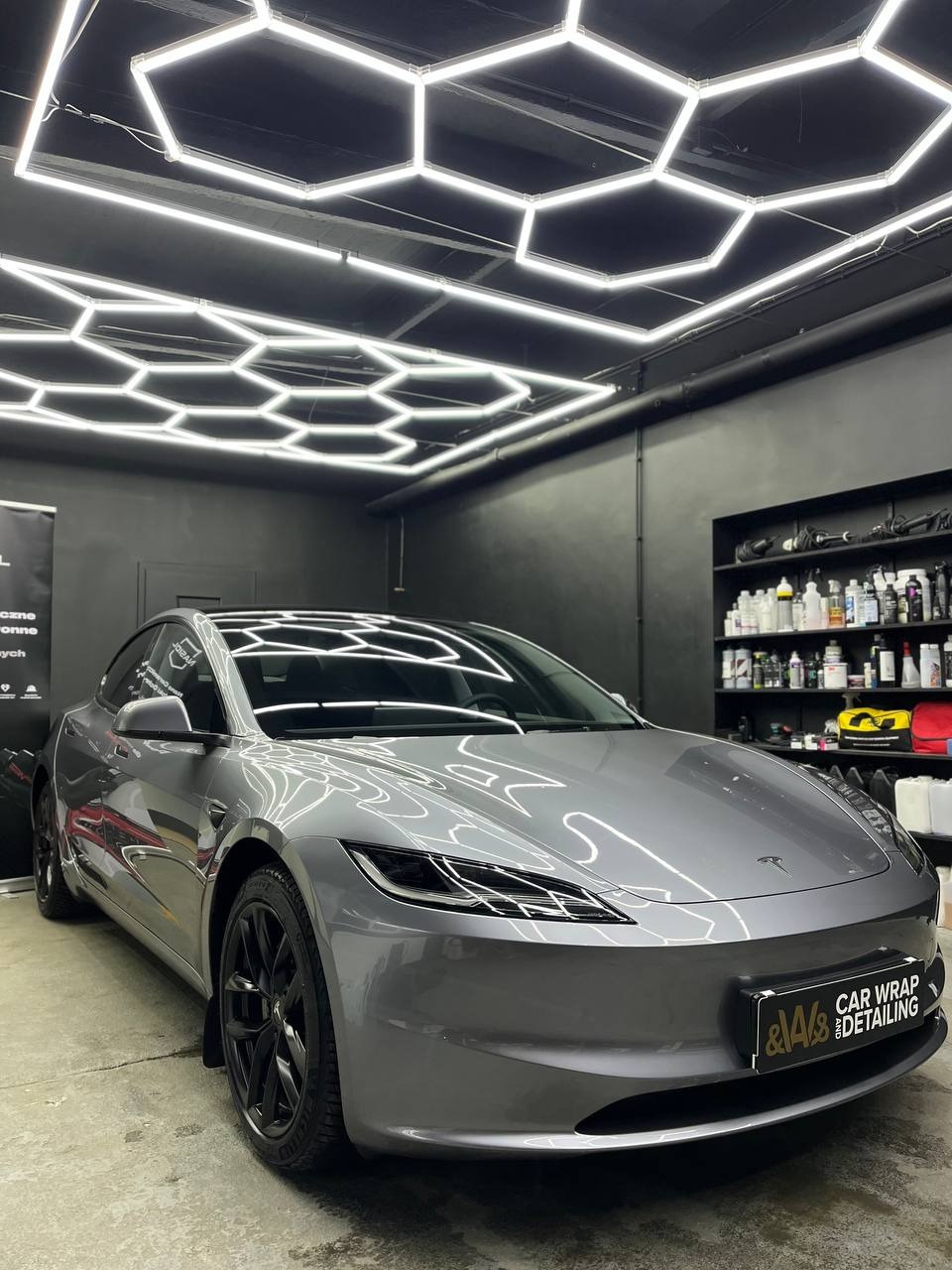 Szary samochód Tesla Model Y po oklejeniu folią w studiu detailingowym z heksagonalnym oświetleniem sufitowym.