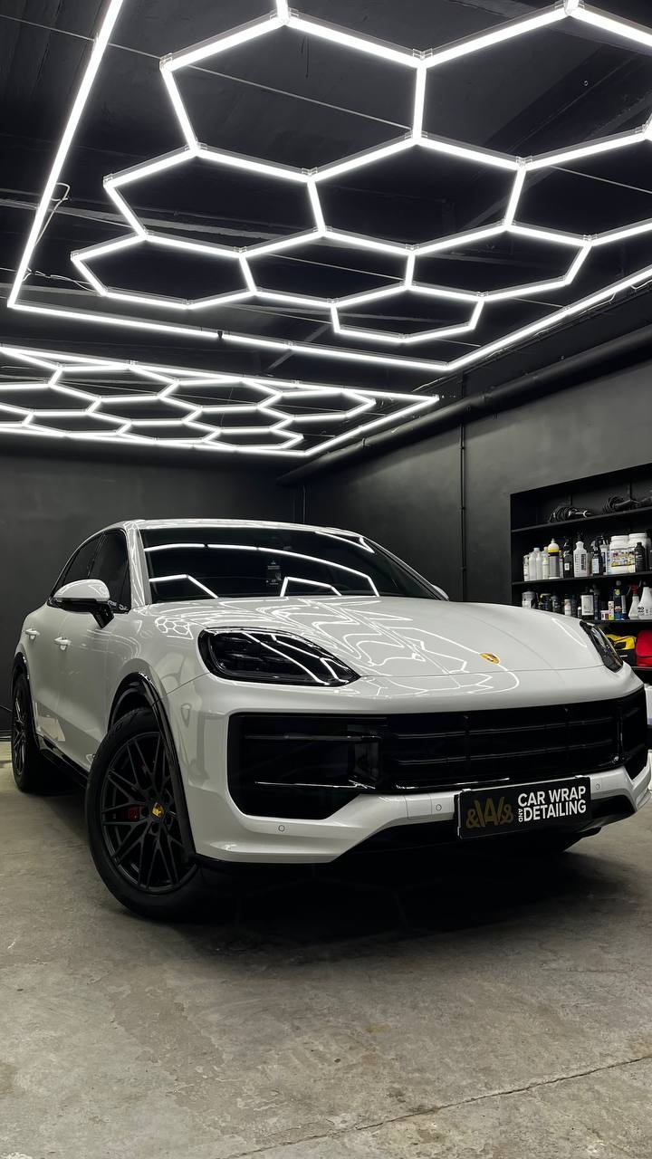 Biały Porsche Cayenne po oklejeniu folią, widoczne logo firmy detailingowej na zderzaku, oświetlenie sufitowe w kształcie plastra miodu.