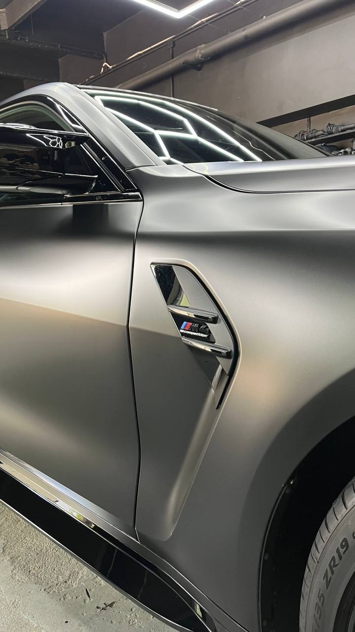 Matowa, szara karoseria BMW M Competition z widocznym emblematem na błotniku, oświetlona studyjnym światłem.