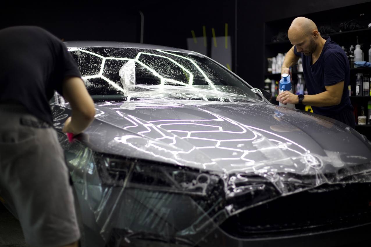 Aplikacja folii ochronnej na przednią część szarego samochodu w warsztacie, widoczni dwaj mężczyźni w trakcie pracy, używający rakli i spryskiwacza.