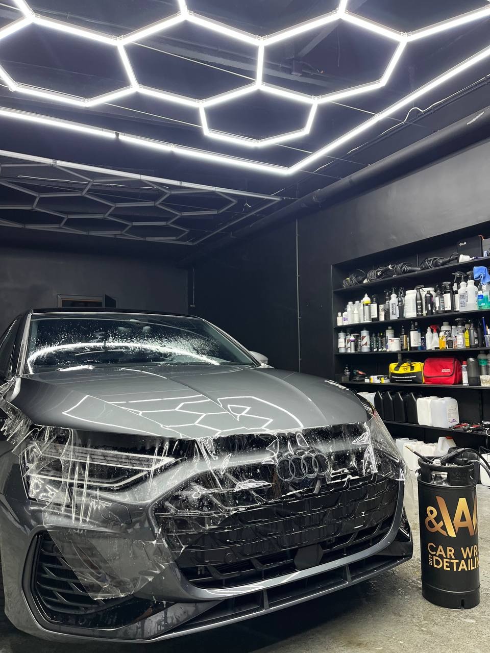 Szary samochód Audi RS7 zabezpieczany folią ochronną w profesjonalnym studiu detailingowym z oświetleniem LED w kształcie plastra miodu i półkami z preparatami do pielęgnacji.