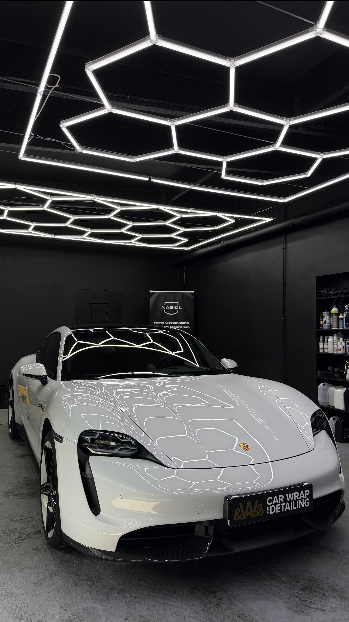 Biały Porsche Taycan w studiu detailingowym, oświetlony heksagonalnymi lampami. Widoczne logo firmy na tablicy rejestracyjnej i kosmetyki na półce w tle.