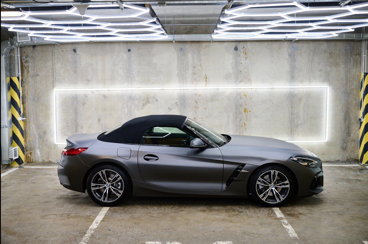Szary, matowy kabriolet BMW Z4 z czarnym dachem, zaparkowany w oświetlonym garażu. Widok z boku, podkreślający detale nadwozia i felgi.