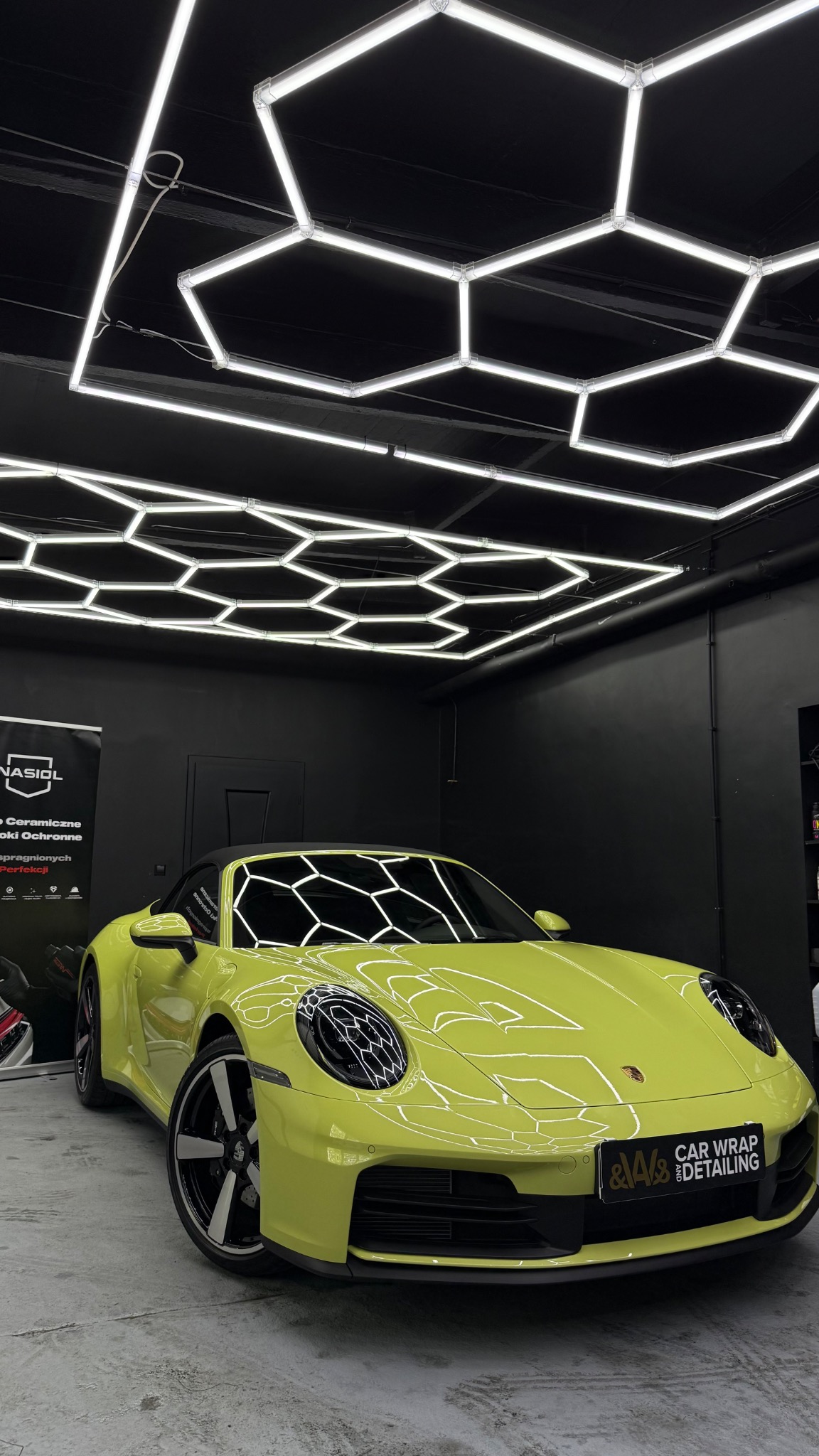 Żółte Porsche 911 po detailingu, lśniące w jasnym studiu z heksagonalnym oświetleniem sufitowym. Widoczne logo firmy detailingowej na przednim zderzaku.