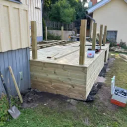 Budowa drewnianego tarasu przy domu jednorodzinnym. Widoczne narzędzia i materiały budowlane, częściowo ukończona konstrukcja, zielony trawnik w tle.