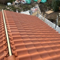 Andrzej Szymański - Ukończony fragment dachu z nową, czerwoną dachówką ceramiczną. Widoczne elementy mocujące i fragment rusztowania w tle. Układanie dachówki w trakcie prac.