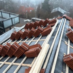 Andrzej Szymański - Układanie brązowej dachówki ceramicznej na drewnianej konstrukcji dachu, widok z góry. Częściowo pokryty membraną dachową, w tle zabudowa jednorodzinna w mglisty dzień.