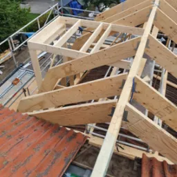 Konstrukcja drewniana dachu w trakcie budowy. Widoczne krokwie, deski poszycia, stara dachówka i rusztowanie. Ujęcie z góry na budowę dachu.