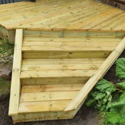 Drewniane schody prowadzące na taras z desek ułożonych pod kątem, widoczne detale konstrukcyjne i naturalne otoczenie z zielenią.