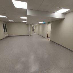 Andrzej Szymański - Jasne, przestronne wnętrze z szarą podłogą, białymi ścianami i sufitami kasetonowymi z wbudowanymi lampami. Widoczne drzwi z zielonymi znakami ewakuacyjnymi.