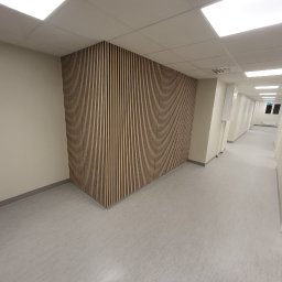 Andrzej Szymański - Panel akustyczny z pionowych listew na ścianie korytarza biurowego. Jasne barwy, minimalistyczny design, równomierne oświetlenie.