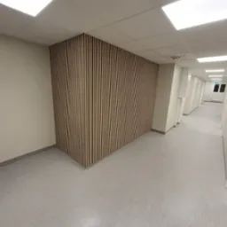 Panel akustyczny z pionowych listew na ścianie korytarza biurowego. Jasne barwy, minimalistyczny design, równomierne oświetlenie.