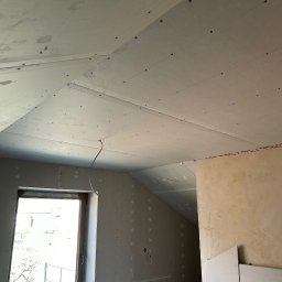 Andrzej Szymański - Poddasze z zamontowanymi płytami karton-gips, widoczne łączenia i śruby. Okno z widokiem na zewnątrz. Częściowo otynkowana ściana w tle. Przewód elektryczny zwisa z sufitu.