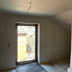 Andrzej Szymański - Poddasze z oknem po montażu płyt kartonowo-gipsowych, widoczne łączenia płyt, przewody elektryczne. Dywan na podłodze zabezpiecza przed zabrudzeniem.