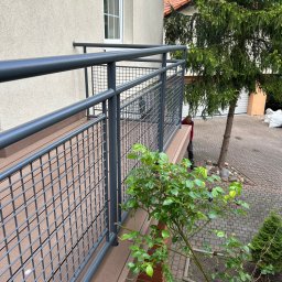 Andrzej Szymański - Nowoczesny balkon z szarą metalową balustradą i brązową deską kompozytową. Widok z góry na roślinność i fragment posesji z kostki brukowej.