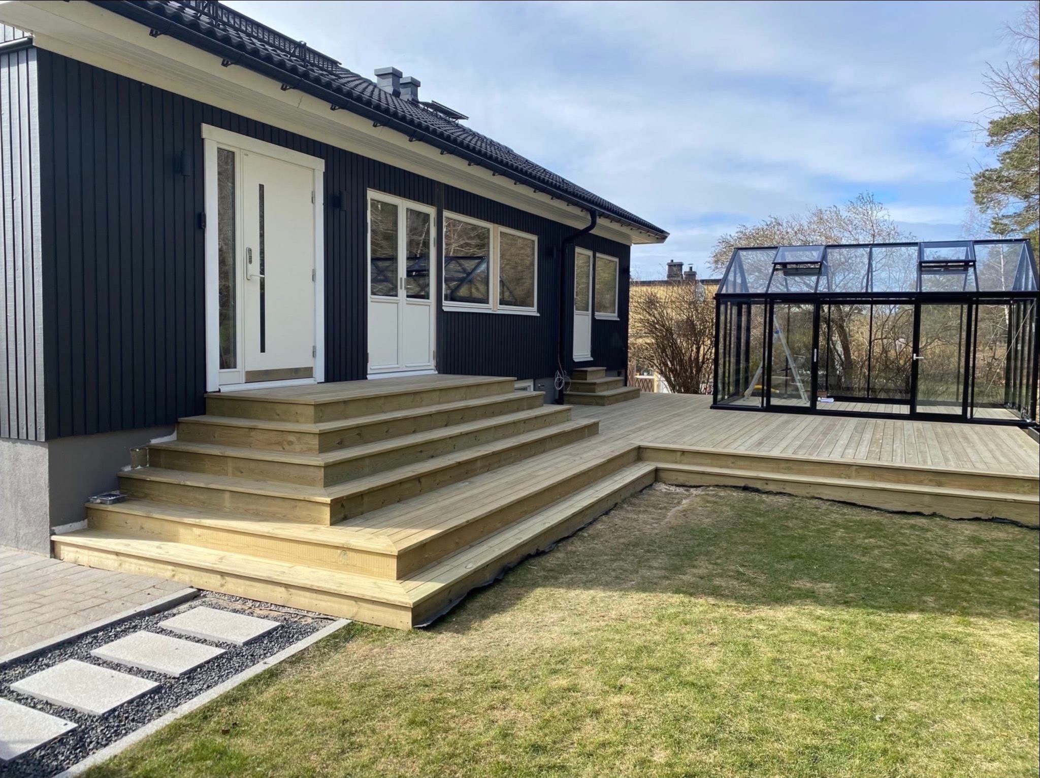 Czarny dom szkieletowy z białymi drzwiami i drewnianym tarasem, na którym stoi szklarnia z czarną ramą. Widok na ogród i fragment ścieżki.