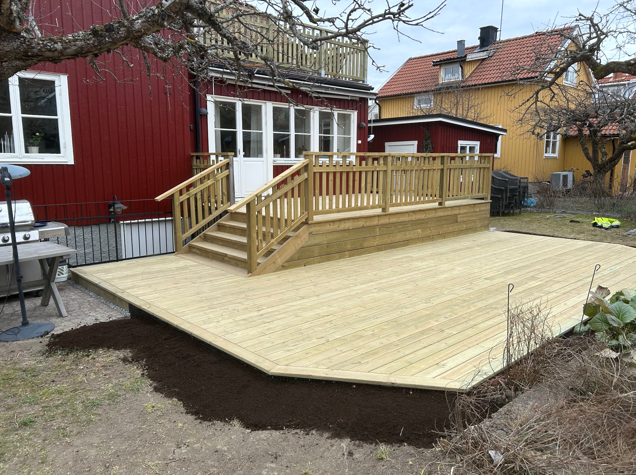 Nowy drewniany taras przy czerwonym domu z białymi oknami i drzwiami. Widoczne schody i barierki. Tło stanowią inne budynki i drzewa.