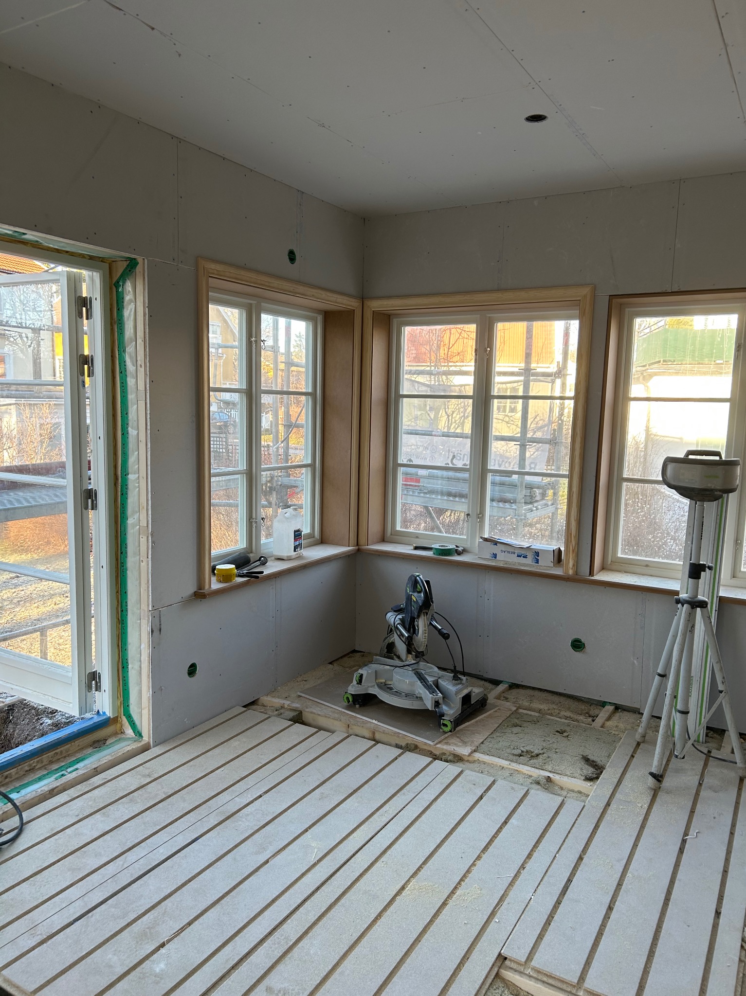 Wnętrze w trakcie wykończenia: płyty gipsowe na ścianach, ułożona podłoga z desek, piła stołowa i statyw oświetleniowy w pokoju z trzema oknami i drzwiami balkonowymi.