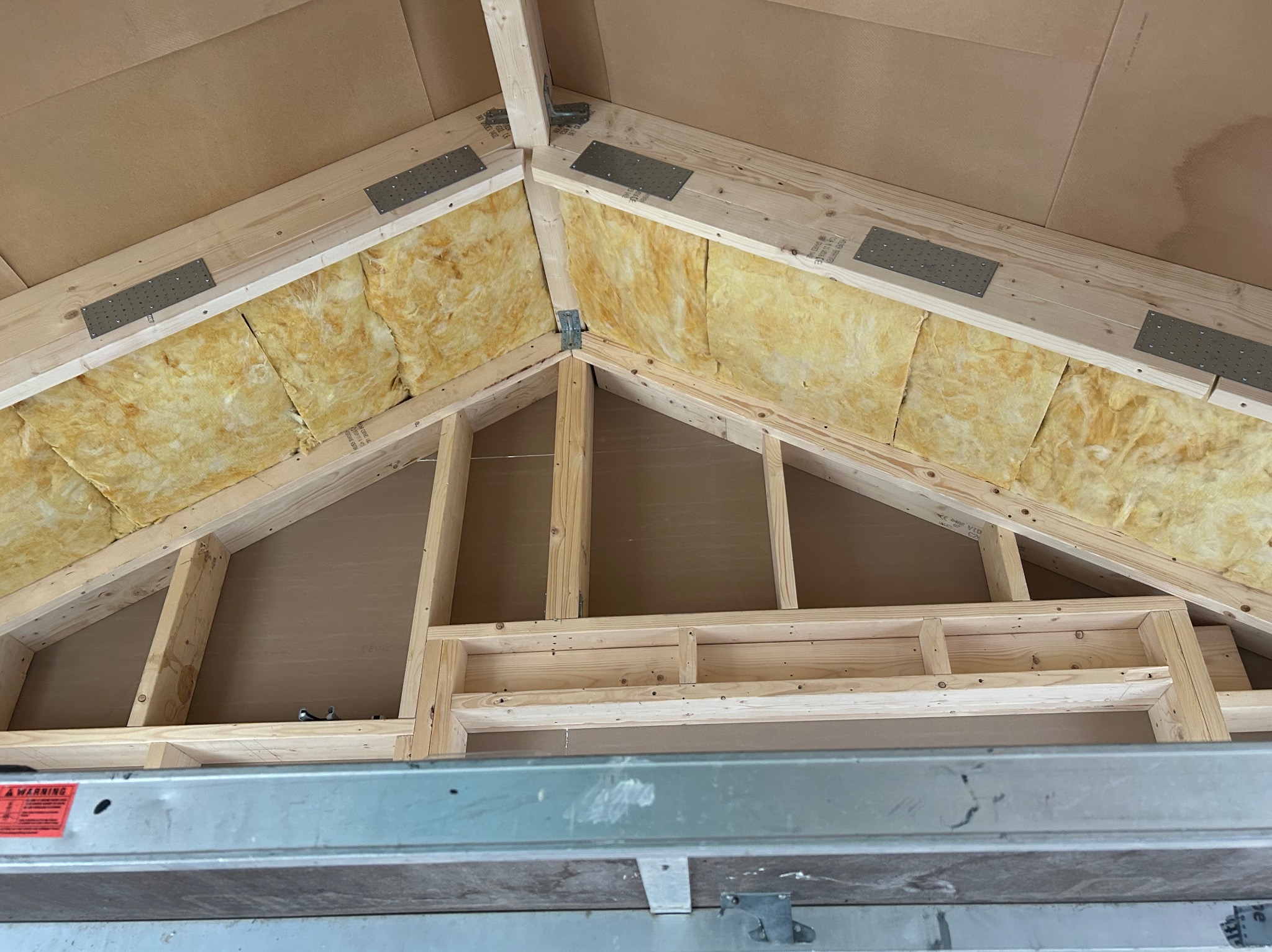 Szkielet dachu z izolacją z wełny mineralnej, widok z dołu. Elementy drewniane połączone metalowymi płytkami. Fragment drabiny w dolnej części kadru.