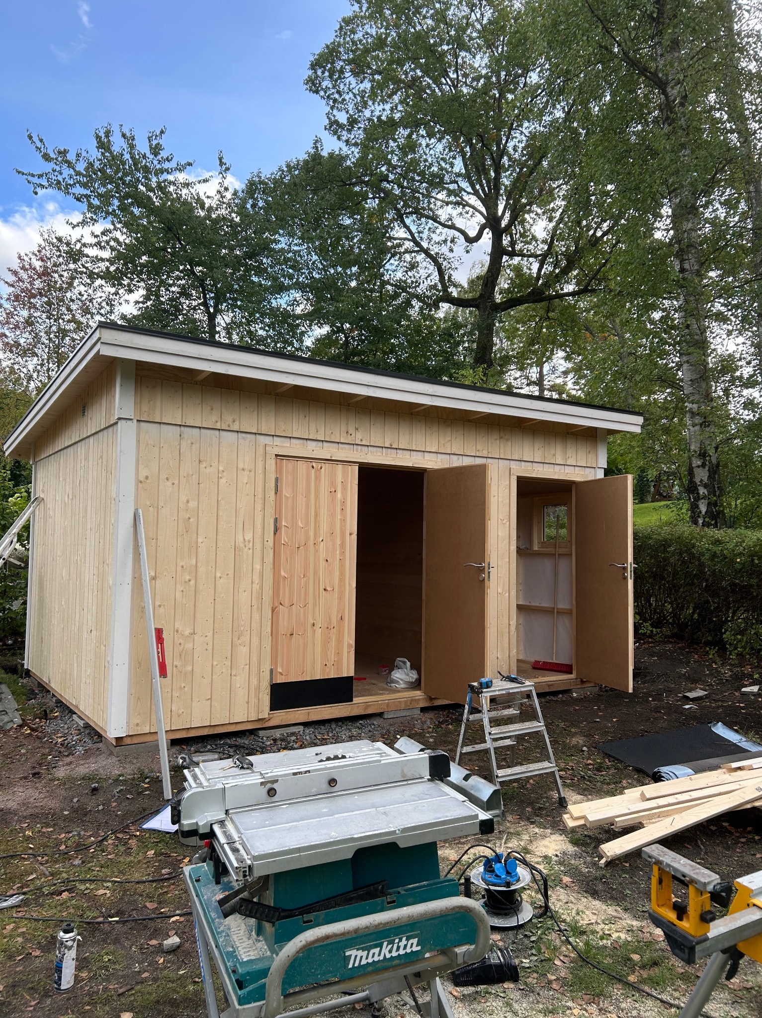 Surowa, drewniana altana z otwartymi drzwiami, ukazująca wnętrze. Widoczne narzędzia i elementy konstrukcyjne na zewnątrz, sugerujące etap budowy w ogrodowym otoczeniu.