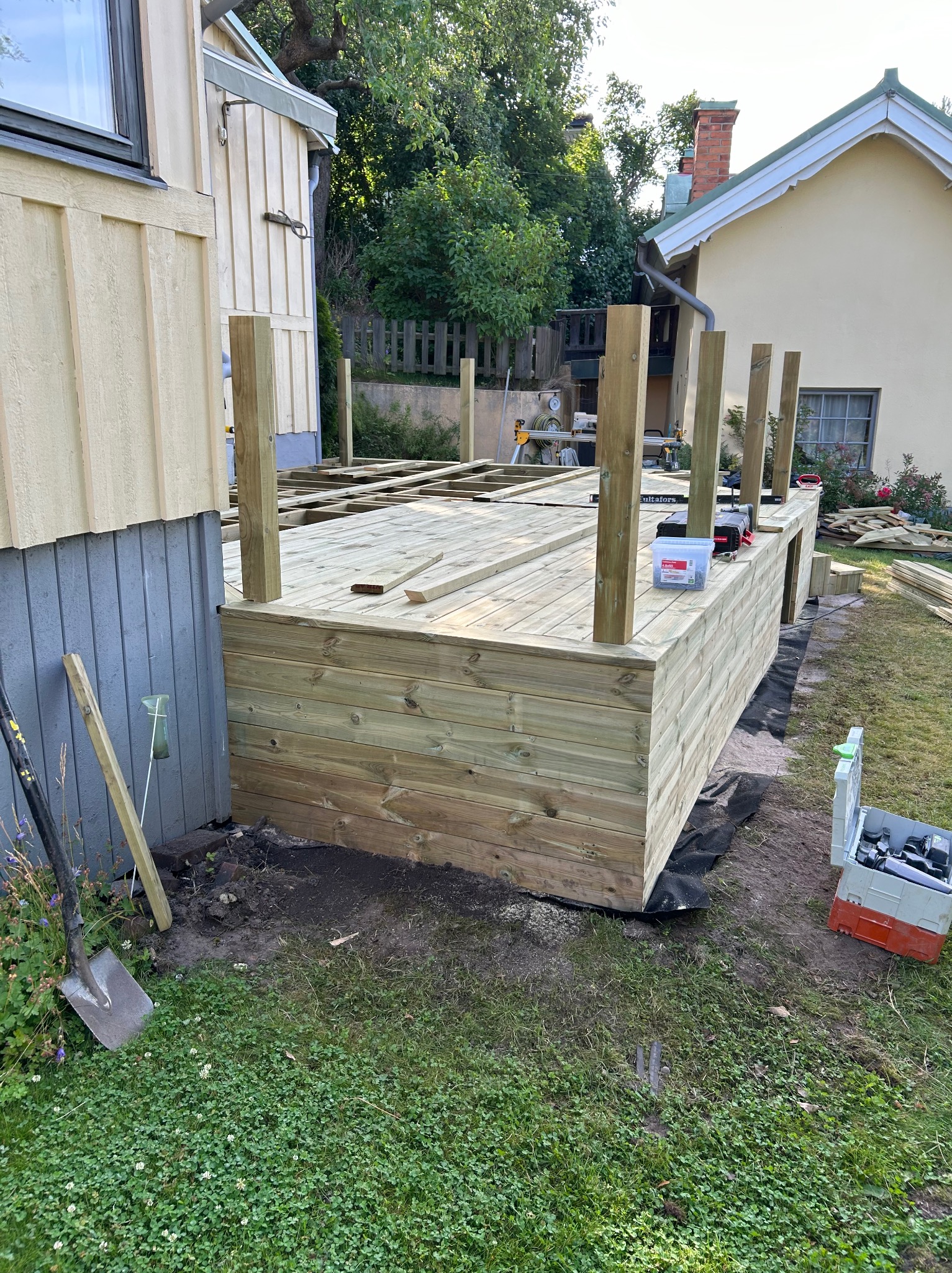 Budowa drewnianego tarasu przy domu jednorodzinnym. Widoczne narzędzia i materiały budowlane, częściowo ukończona konstrukcja, zielony trawnik w tle.