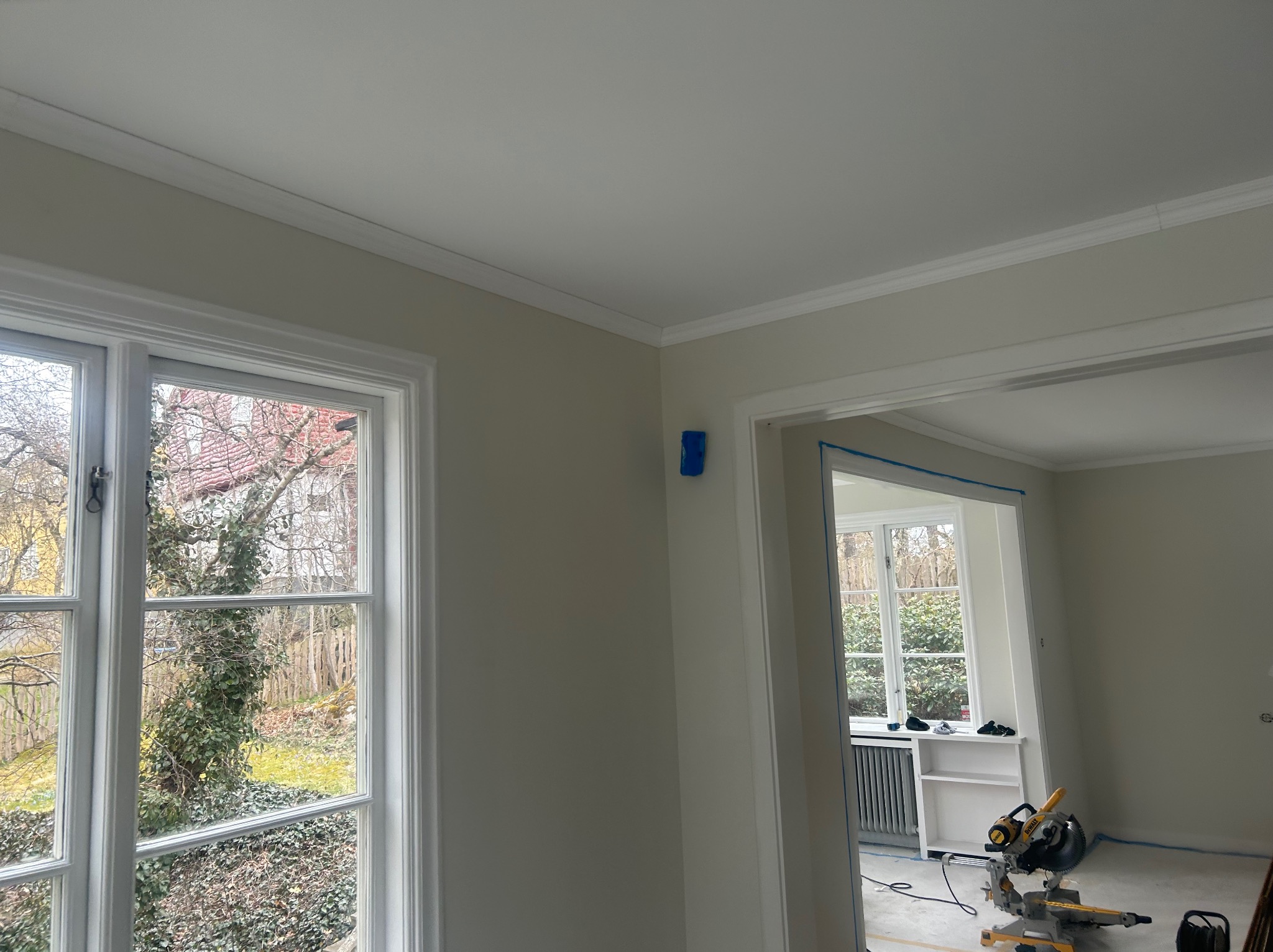Jasne wnętrze w trakcie wykańczania: świeżo pomalowane ściany, listwy przysufitowe, okno z widokiem na ogród, piła do cięcia paneli pod ścianą.