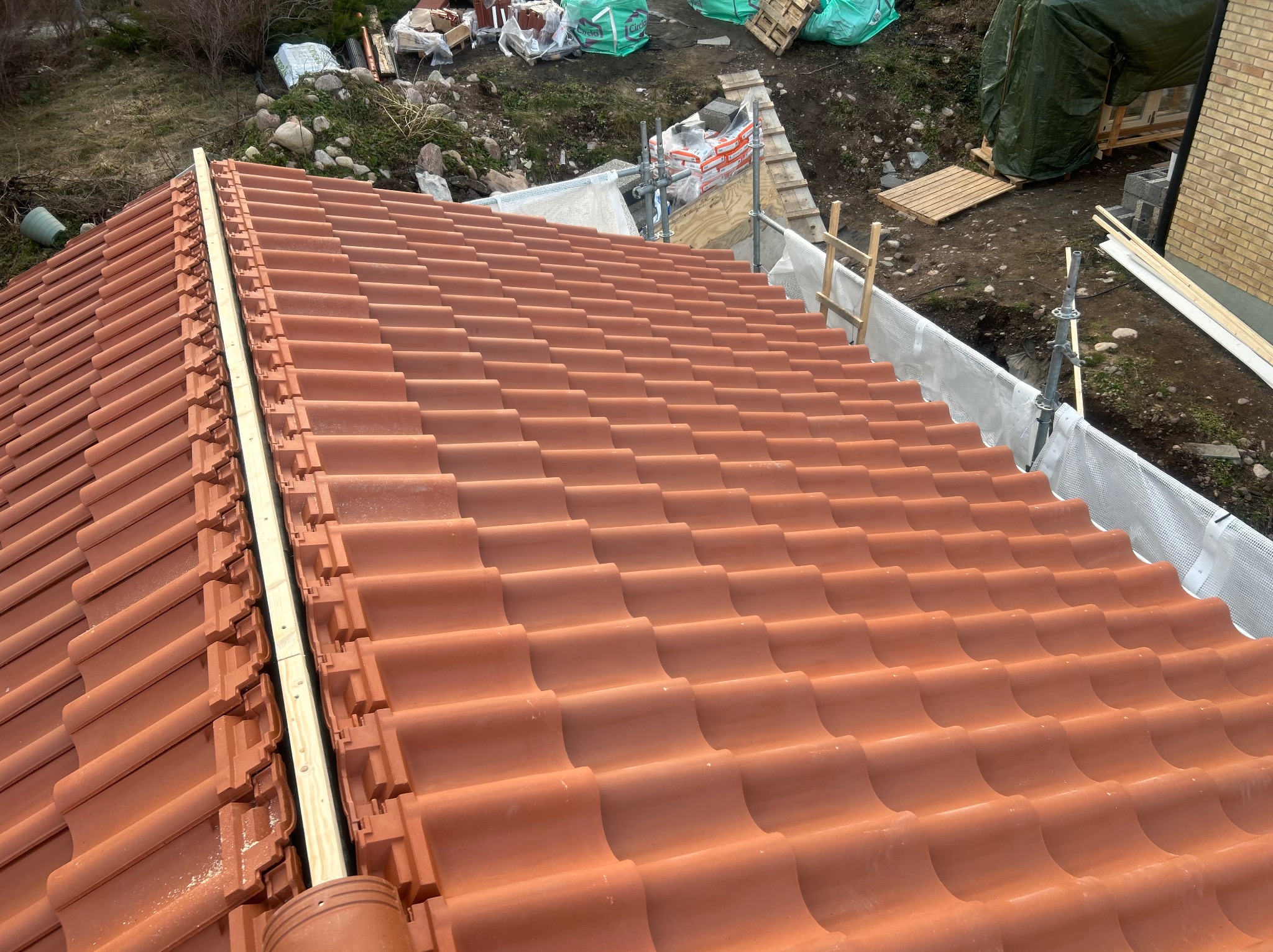 Ukończony fragment dachu z nową, czerwoną dachówką ceramiczną. Widoczne elementy mocujące i fragment rusztowania w tle. Układanie dachówki w trakcie prac.