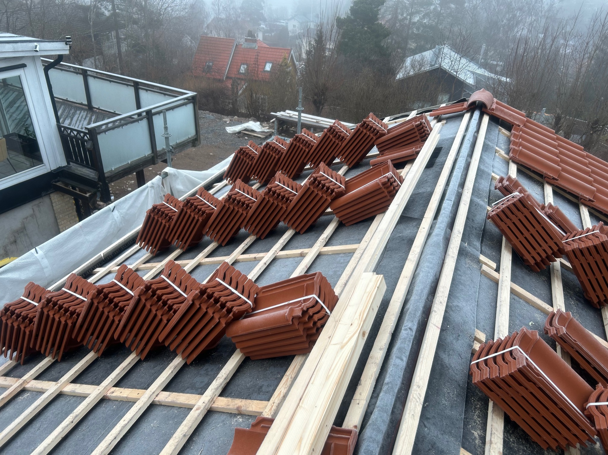 Układanie brązowej dachówki ceramicznej na drewnianej konstrukcji dachu, widok z góry. Częściowo pokryty membraną dachową, w tle zabudowa jednorodzinna w mglisty dzień.