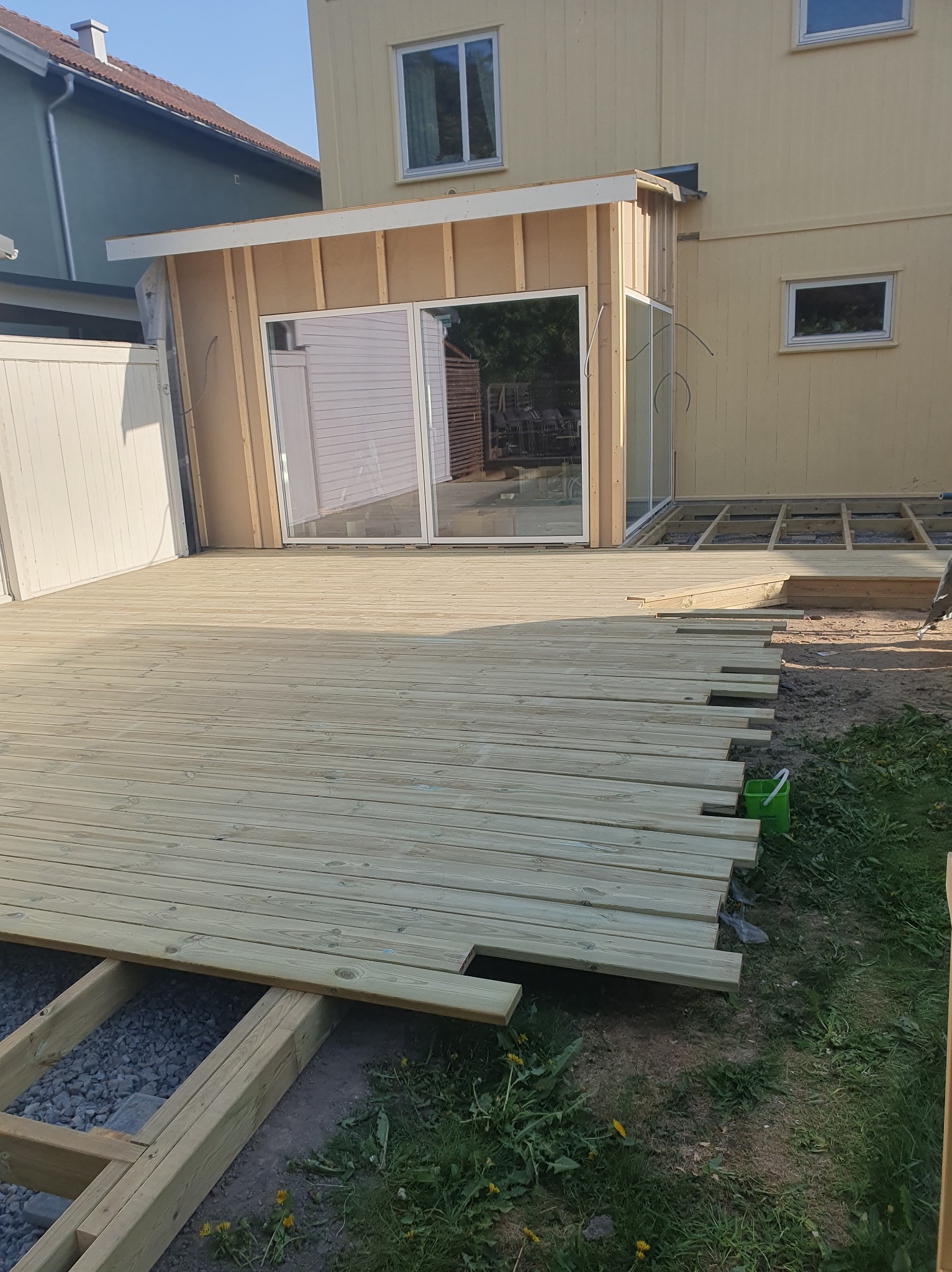 Jasna, drewniana deska tarasowa w trakcie montażu przy domu z dużym oknem przesuwnym. Część desek jeszcze nieułożona, widoczna konstrukcja pod spodem.