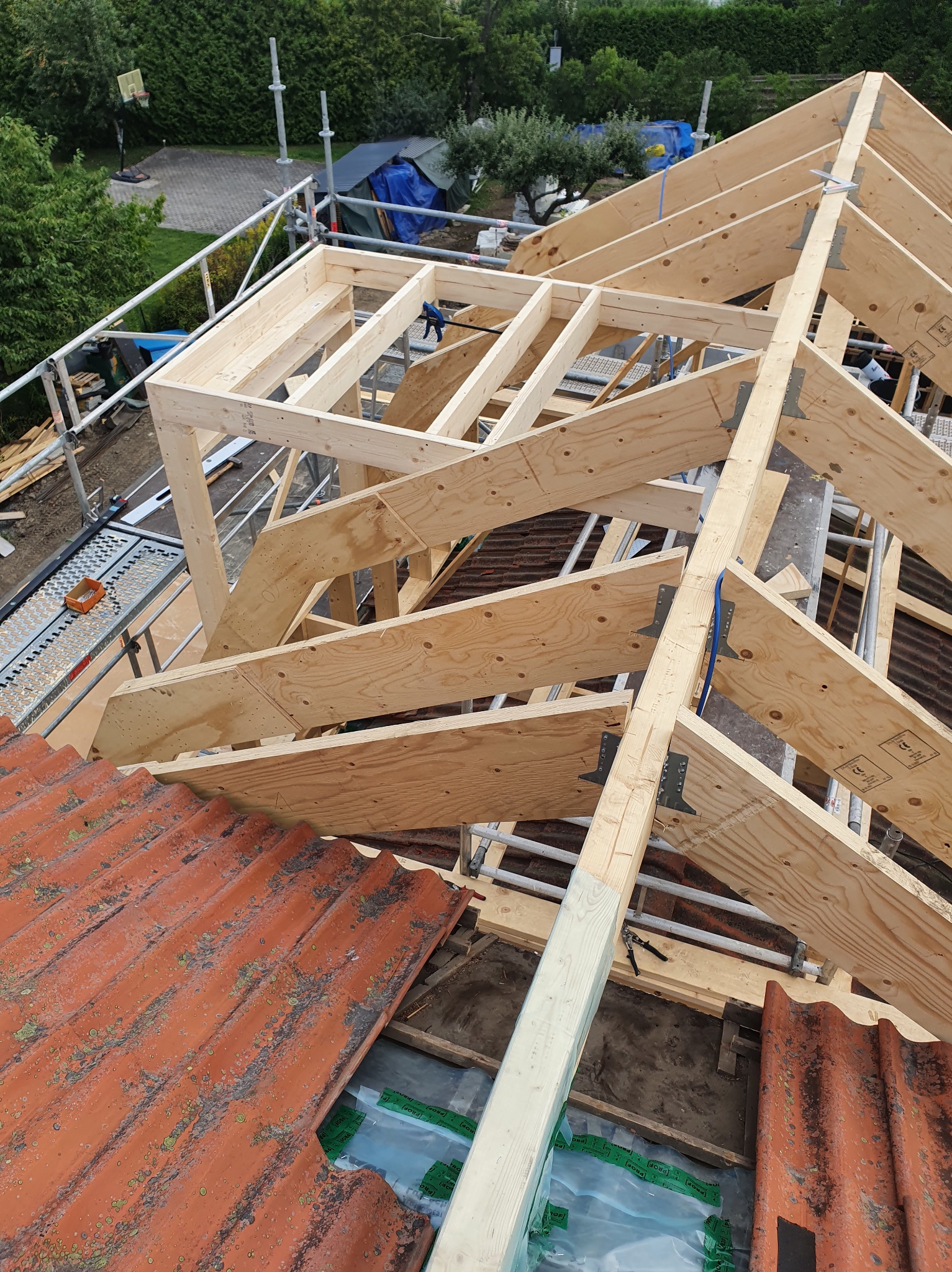 Konstrukcja drewniana dachu w trakcie budowy. Widoczne krokwie, deski poszycia, stara dachówka i rusztowanie. Ujęcie z góry na budowę dachu.