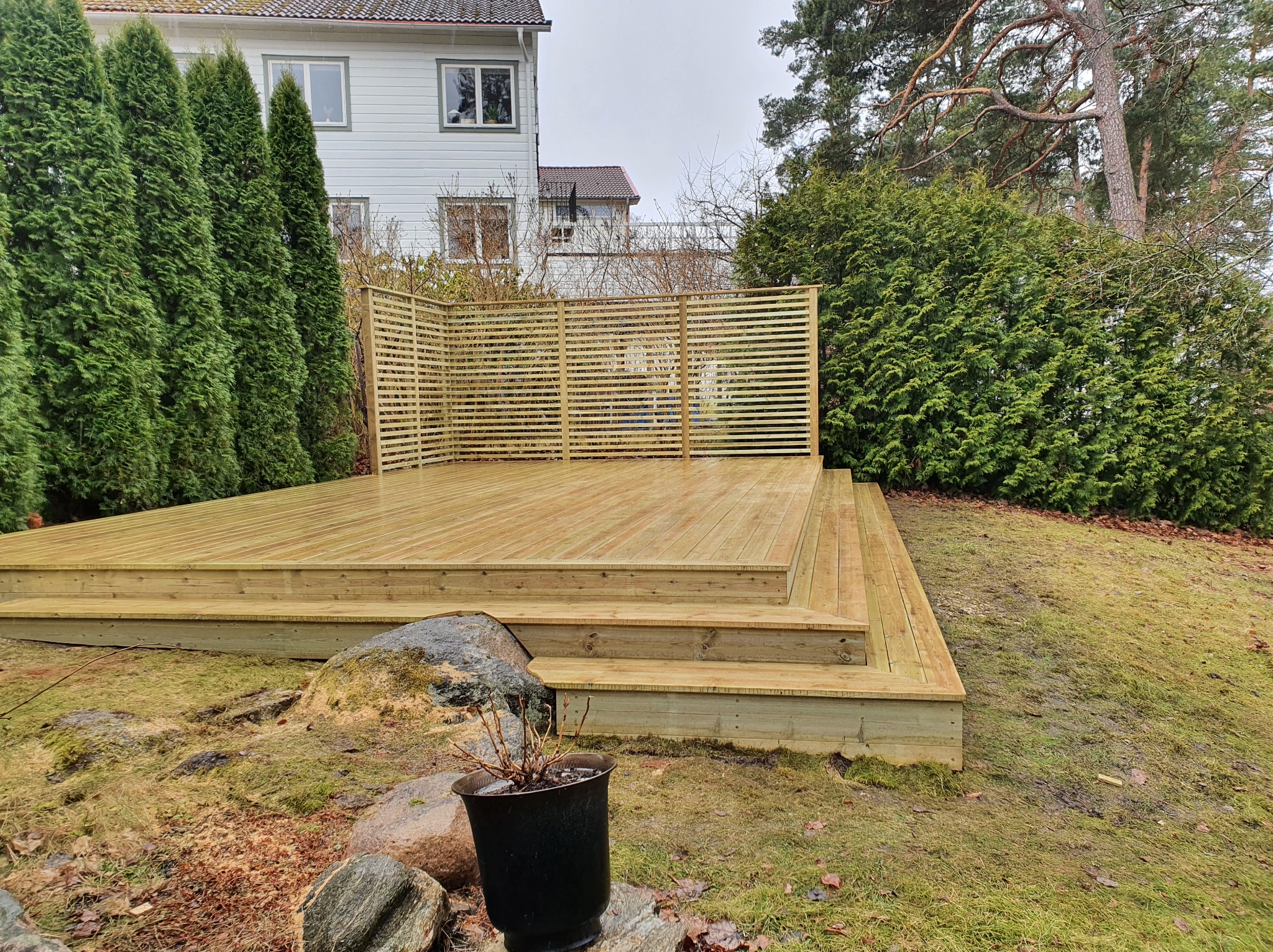 Nowoczesny taras drewniany z ażurową ścianką, usytuowany w ogrodzie z zielenią i kamieniami, w tle dom. Świeżo zaimpregnowane deski tworzą jasny, naturalny wygląd.