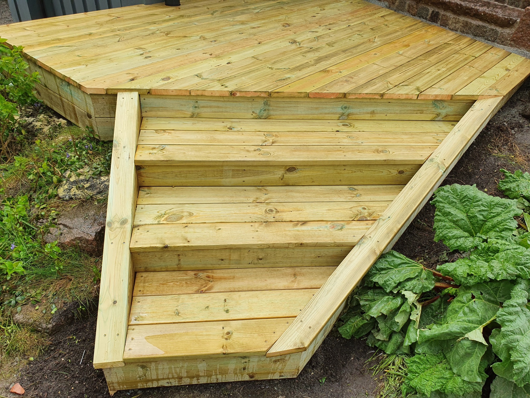 Drewniane schody prowadzące na taras z desek ułożonych pod kątem, widoczne detale konstrukcyjne i naturalne otoczenie z zielenią.