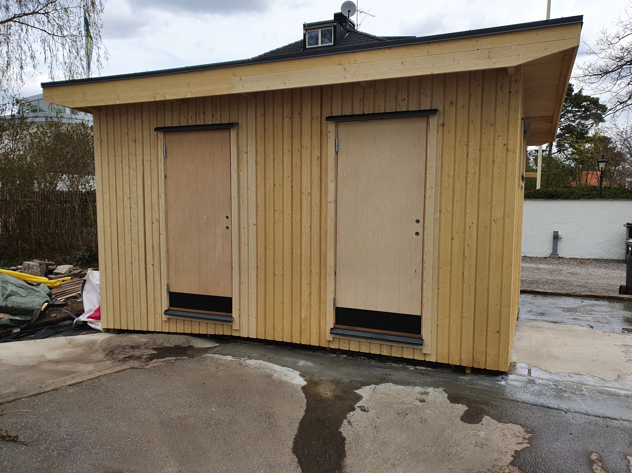 Nowoczesna, drewniana konstrukcja z dwoma jasnymi drzwiami i czarnymi detalami, na tle zieleni i szarej nawierzchni, w pochmurny dzień.