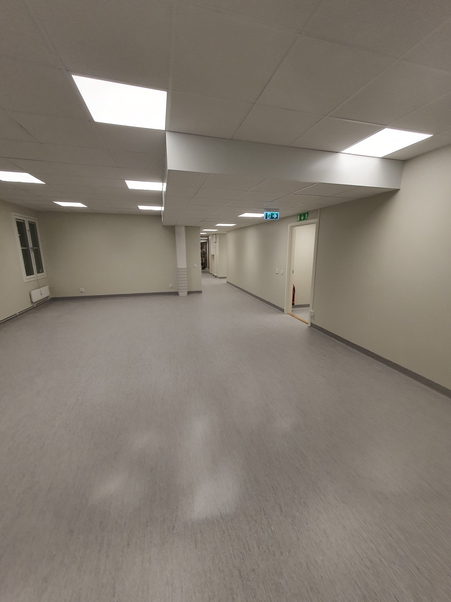 Jasne, przestronne wnętrze z szarą podłogą, białymi ścianami i sufitami kasetonowymi z wbudowanymi lampami. Widoczne drzwi z zielonymi znakami ewakuacyjnymi.