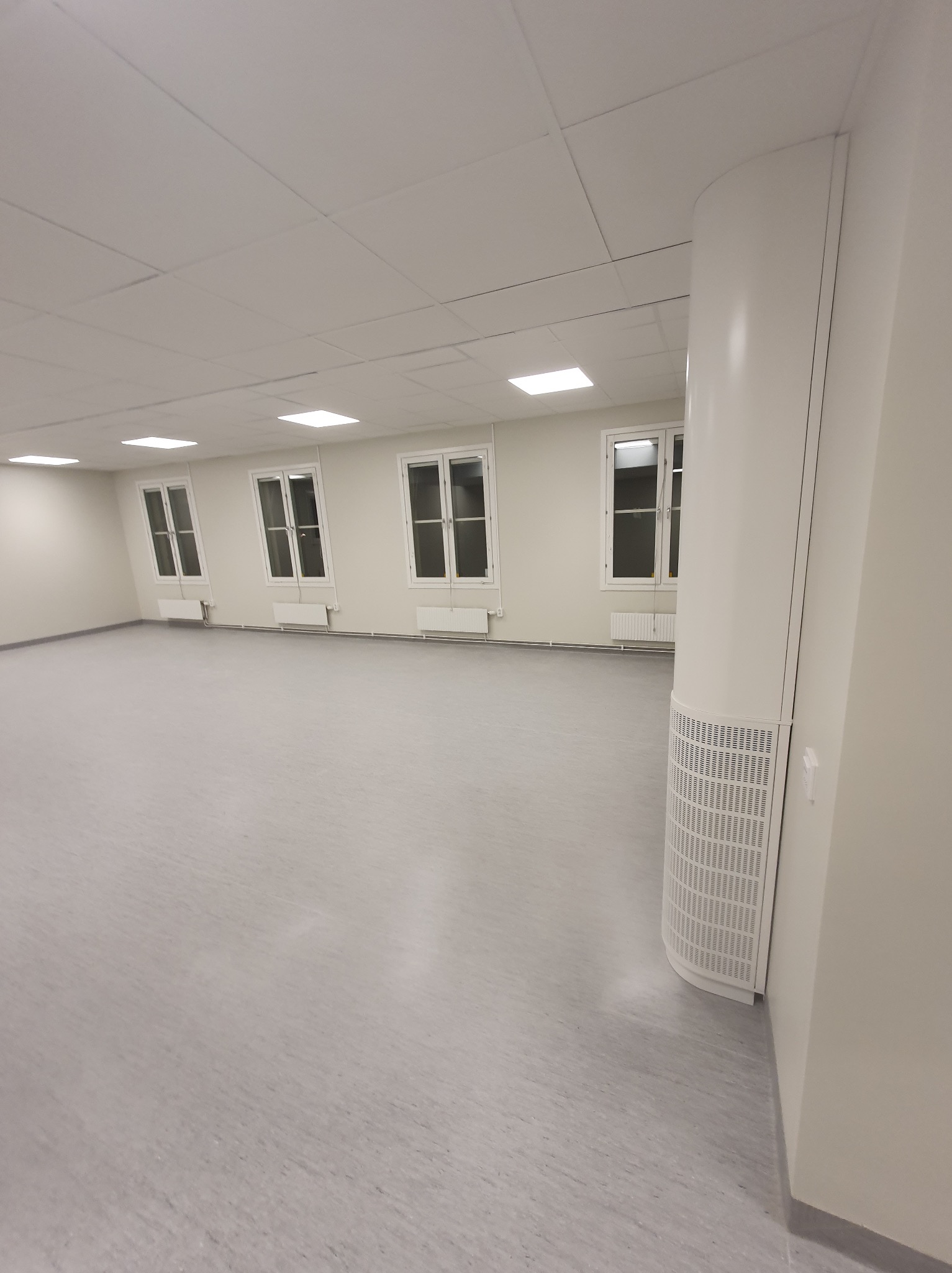 Jasne, puste wnętrze z szarym linoleum, białymi ścianami i oknami. Widoczny fragment kolumny wentylacyjnej. Nowoczesne wykończenie.
