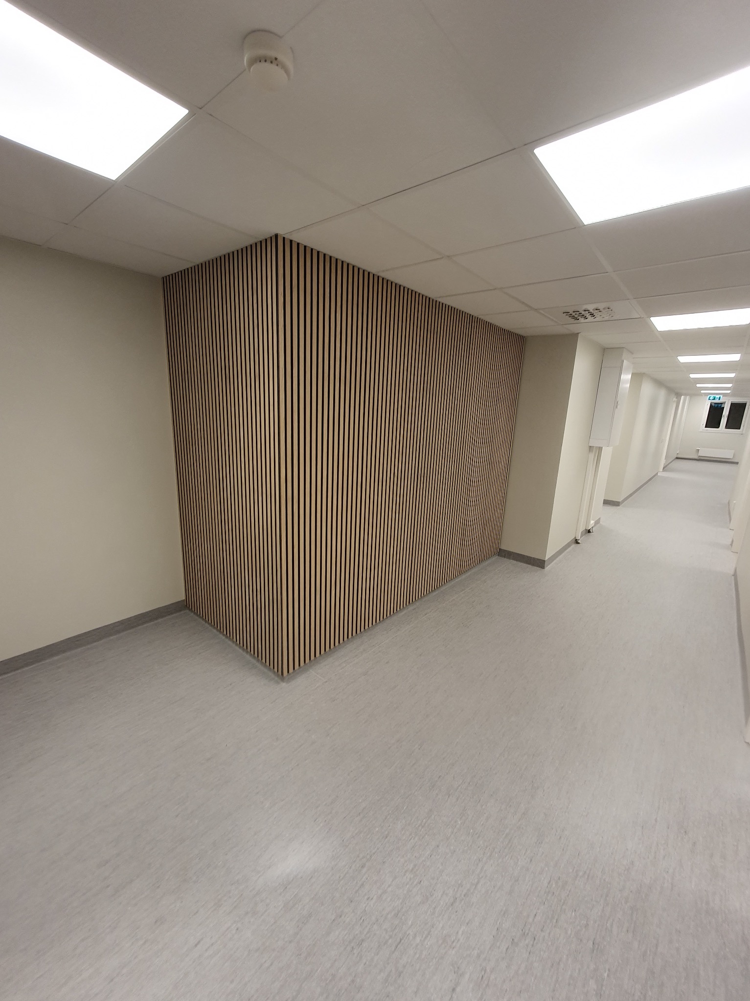 Panel akustyczny z pionowych listew na ścianie korytarza biurowego. Jasne barwy, minimalistyczny design, równomierne oświetlenie.