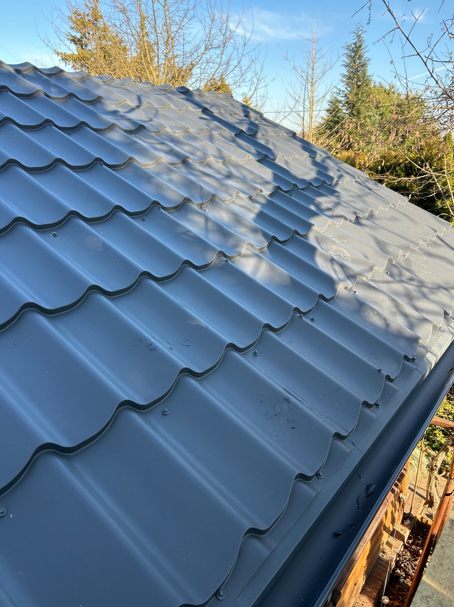 Szary dach z blachy trapezowej, widok z góry, z widocznymi drzewami w tle i elementami systemu rynnowego. Nowoczesne pokrycie dachowe.