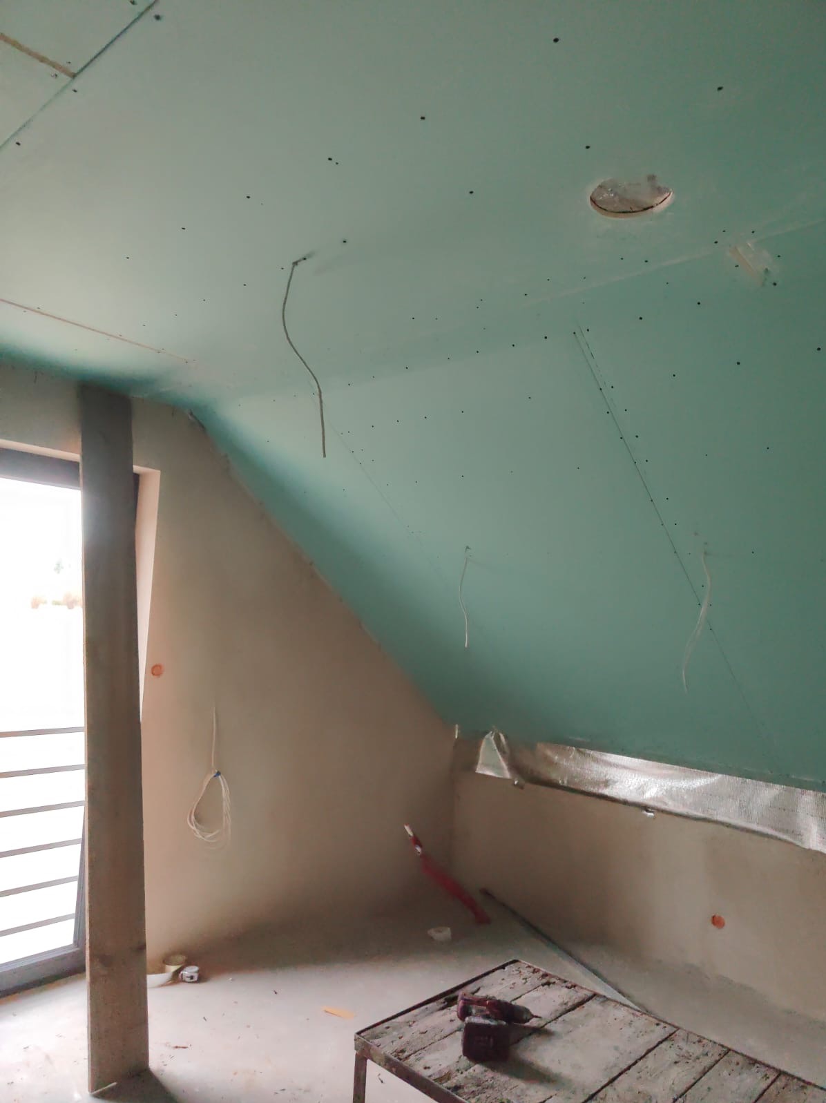 Skośne poddasze z zamontowaną zieloną płytą karton-gips, widoczne kable i narzędzia na stole. Etap prac wykończeniowych w pomieszczeniu ze skosami i oknem.