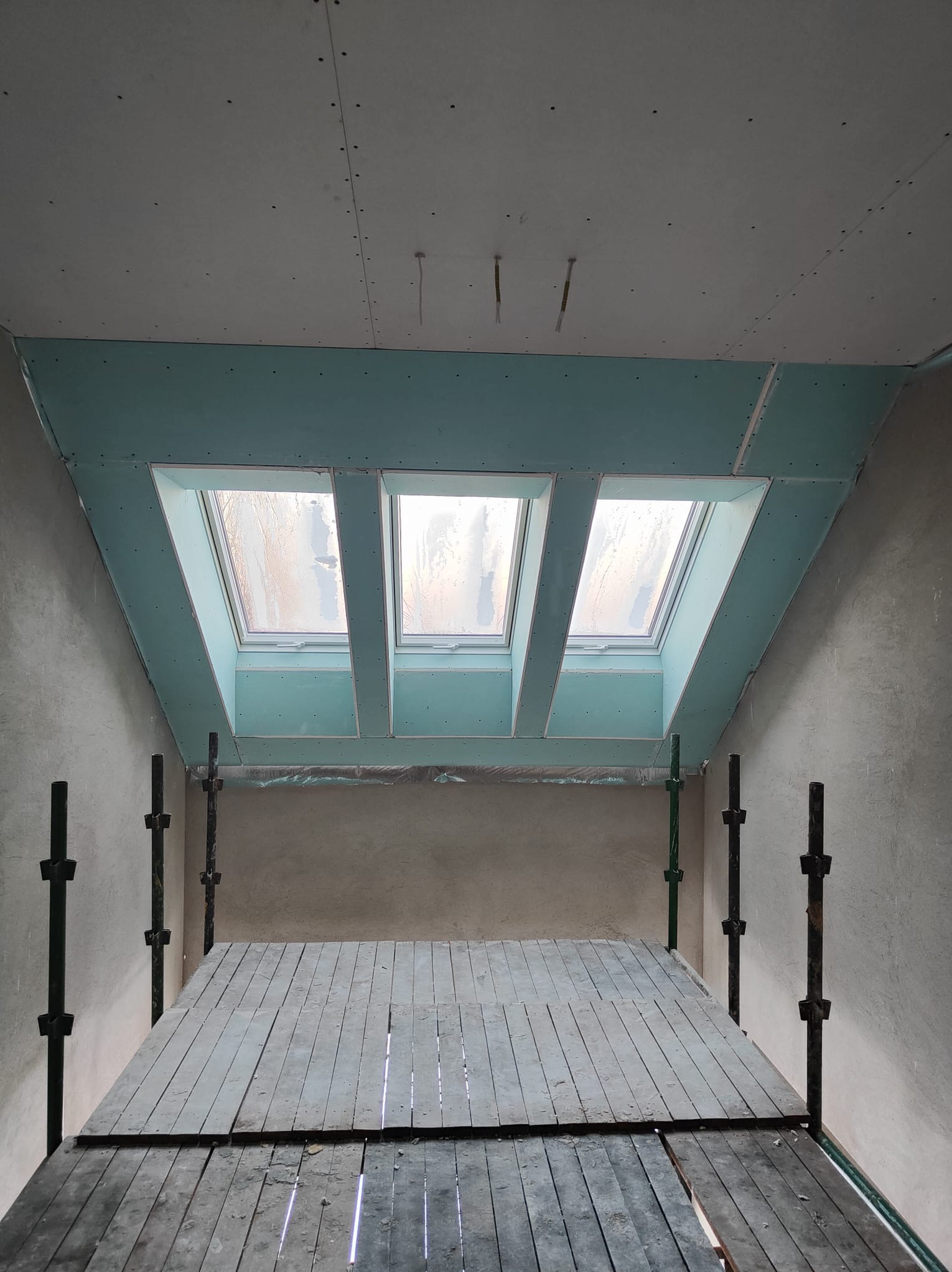 Montaż płyt G-K na poddaszu z oknami dachowymi, widoczne rusztowanie. Szpachlowane łączenia płyt, surowy stan ścian. Ujęcie z dołu, podkreślające skos dachu.